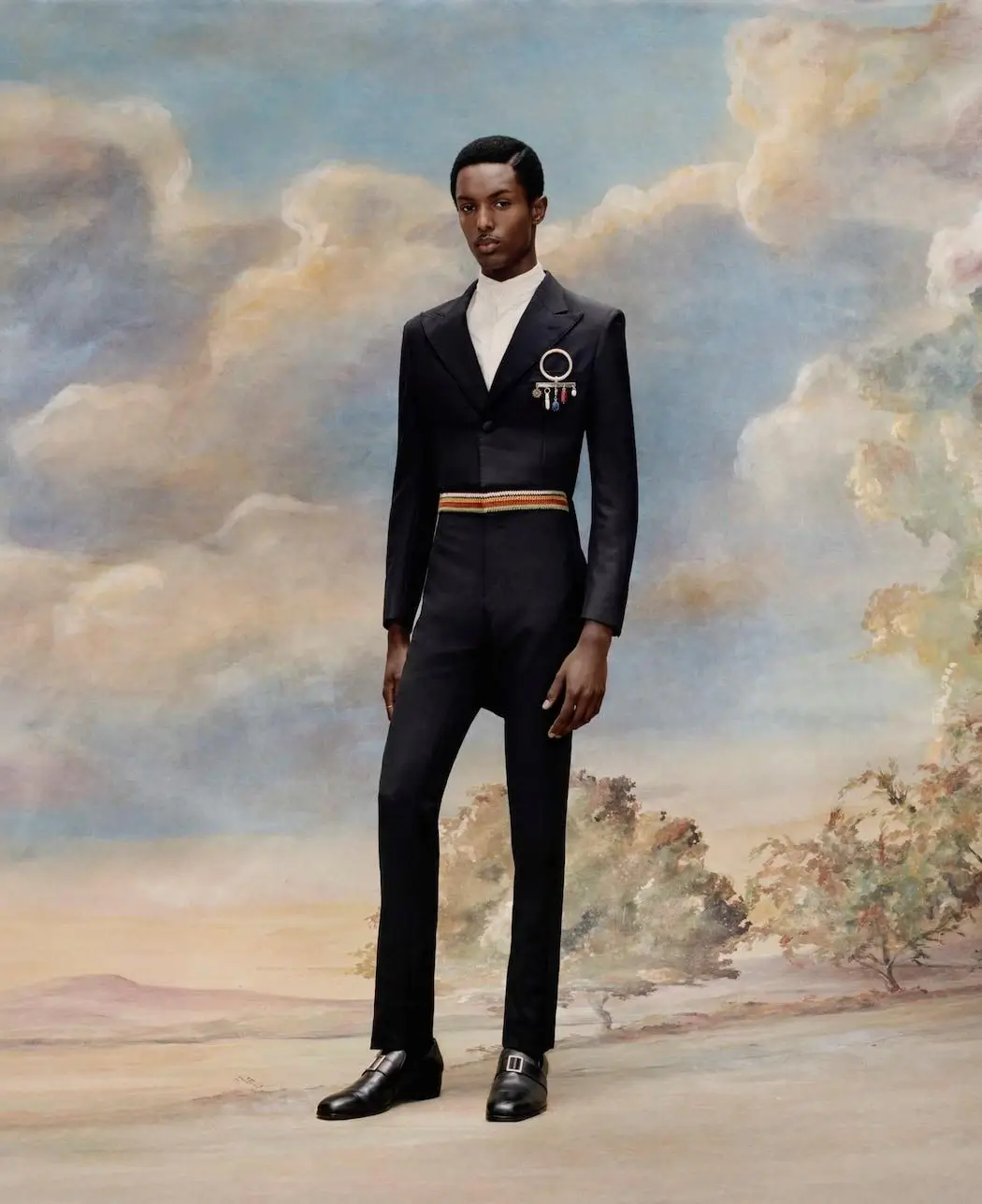 Model posed against a painted, pastoral sky backdrop with soft clouds and landscape details, wearing a tailored black suit with a fitted silhouette and slightly cropped trousers. The look is paired with a high-collared white shirt and a decorative brooch pinned to the chest, featuring a circular ring with hanging embellishments. A striped waistband detail adds contrast at the midsection, while polished black leather shoes complete the refined, formal ensembl