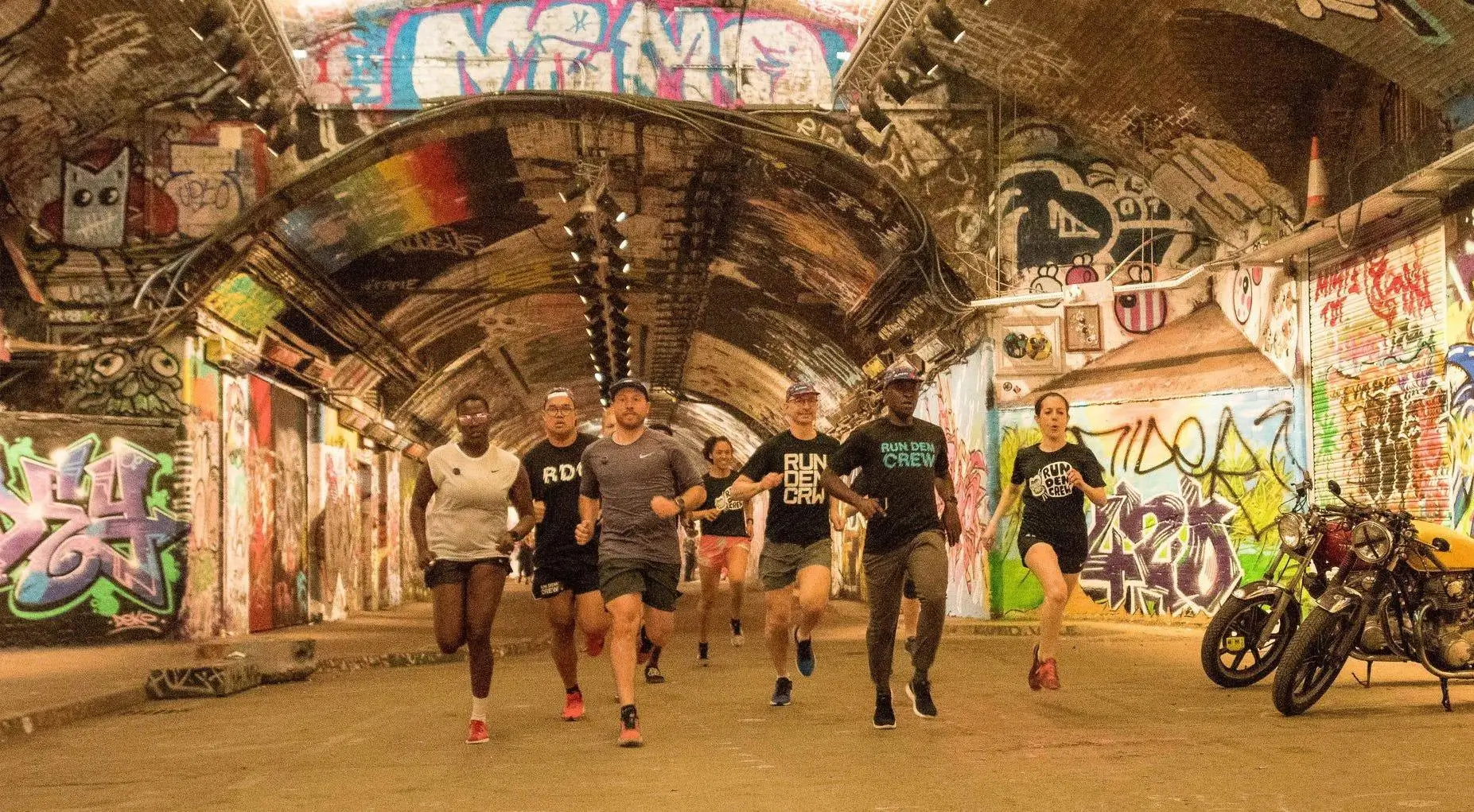 A diverse group of runners jogs through a graffiti-covered urban tunnel, wearing athletic gear with “Run Dem Crew” branding, surrounded by colorful street art, overhead lights, and parked motorcycles along the side
