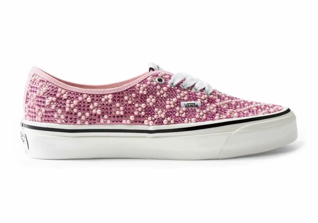 Side profile of pink Vans Authentic shoe fully covered in rhinestones and pearl-like studs, featuring white laces, a small Vans tag, and an off-white rubber sole against a plain background