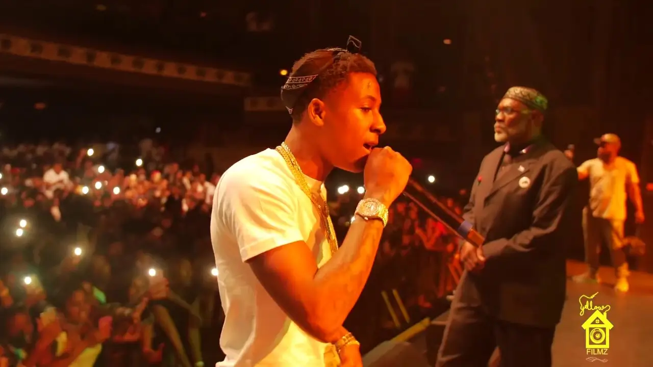 Rapper performing on stage holding a microphone, wearing a white t-shirt, gold chains, and a watch, with a large crowd behind him holding up phones while another man stands nearby under warm concert lighting