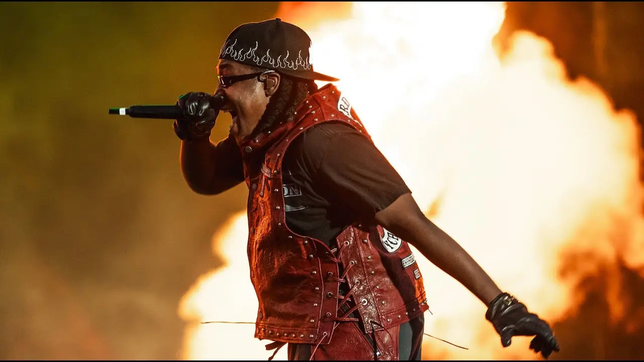 Performer on stage mid-set, wearing a red studded vest, black cap with flame detailing, and gloves, holding a microphone while rapping in front of a massive burst of pyrotechnic flames
