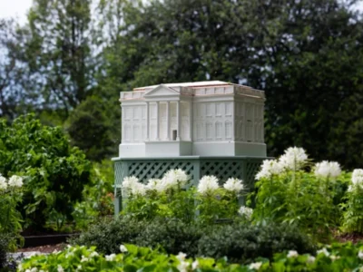 Miniature White House–shaped beehive set on a raised platform within the South Lawn Kitchen Garden, surrounded by blooming white flowers and lush greenery, highlighting its detailed architectural design and role in pollination