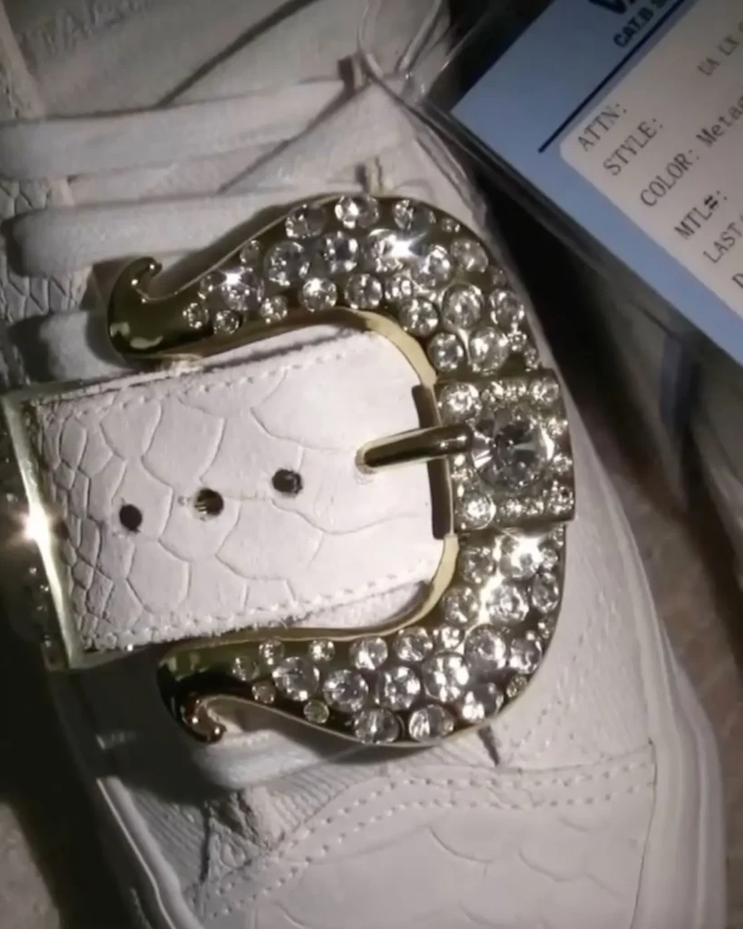 A close-up detail of a white leather accessory—likely a belt or handbag strap—featuring an ornate, oversized gold-tone buckle encrusted with clustered, multi-sized clear rhinestones. The leather has a subtle embossed texture with visible stitching, while the buckle curves into a sculptural, almost baroque silhouette. Light catches the stones, creating sharp, reflective highlights that contrast against the matte grain of the leather. A product tag sits partially in frame, suggesting retail or showroom context