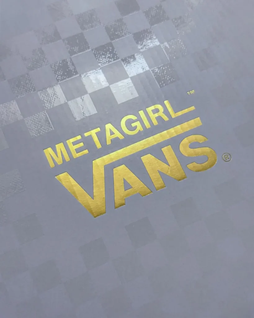 A close-up of branded packaging featuring a tonal grey checkerboard background with alternating matte and glossy squares, subtly catching the light. Centered across the surface is a bold, metallic gold logo reading “METAGIRL VANS®,” rendered in a clean, sans-serif typeface with the iconic Vans underline extending from the “V.” The foil-stamped lettering reflects light with a polished sheen, creating contrast against the muted, textured backdrop. The overall presentation feels premium and collaborative, hinting at a special-edition release or co-branded product