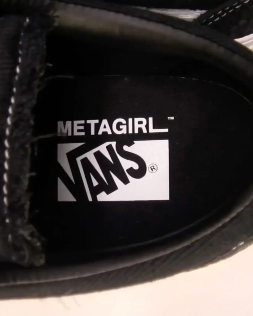 A tight interior shot of a black shoe reveals co-branded labeling on the insole. The surface features a textured, mesh-like fabric, overlaid with a crisp white graphic: “METAGIRL™” sits above the classic Vans logo, which is partially tilted within a rectangular frame, maintaining its signature skate aesthetic. The monochrome contrast—white print against deep black—feels sharp and utilitarian, while the stitching and padded edges around the collar emphasize durability and structure. The composition underscores a collaborative identity, merging Metagirl’s branding with Vans’ iconic visual language in a clean, embedded finish