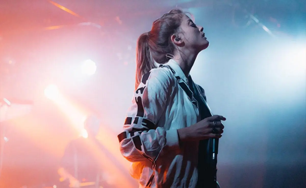 Noga Erez performing live on stage, captured in a moment of intensity with eyes closed and head tilted upward, illuminated by contrasting red and blue stage lights against a hazy concert backdrop