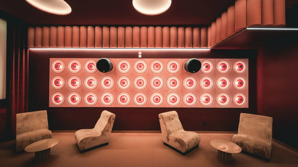 A warmly lit cinema lounge interior features sculptural seating and a wall of circular acoustic panels glowing in soft red tones, creating an immersive, design-forward viewing atmosphere