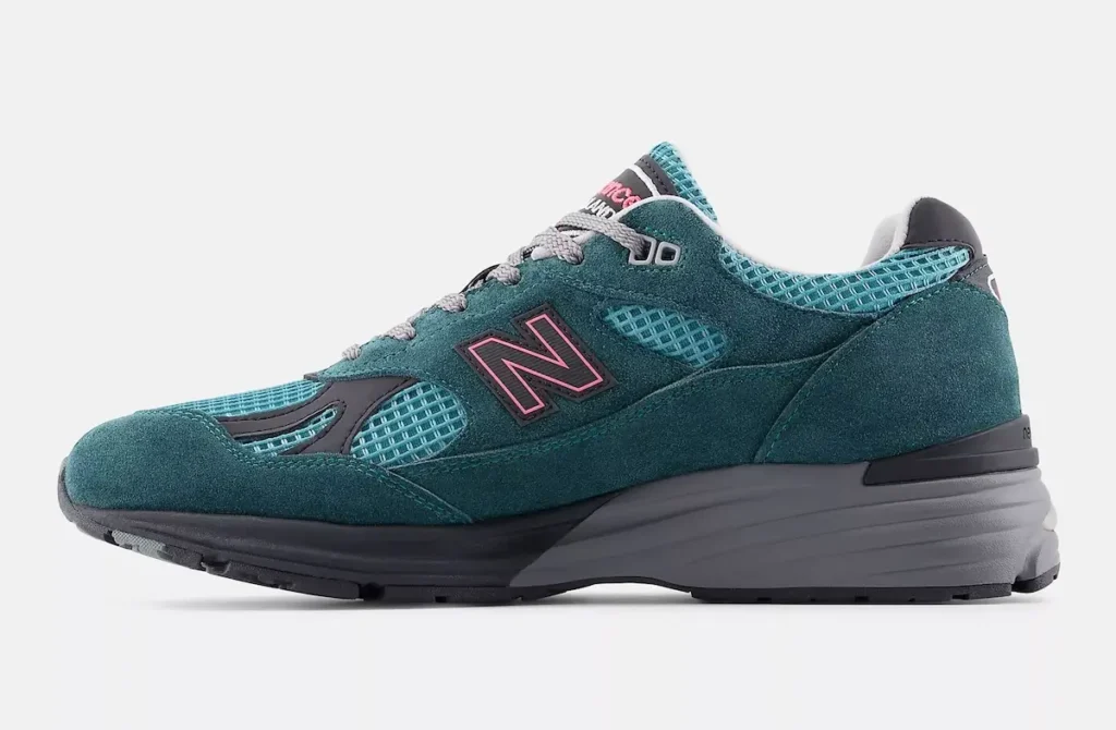 Side-profile studio image of a New Balance 991v2 shoe in a teal and aqua colorway, combining soft suede overlays with breathable mesh panels. The shoe features grey laces, a sculpted dark midsole, and a black outsole, with pink accents outlining the signature “N” logo for contrast. The clean lateral view highlights the streamlined silhouette, layered construction, and balance between performance heritage and modern lifestyle design