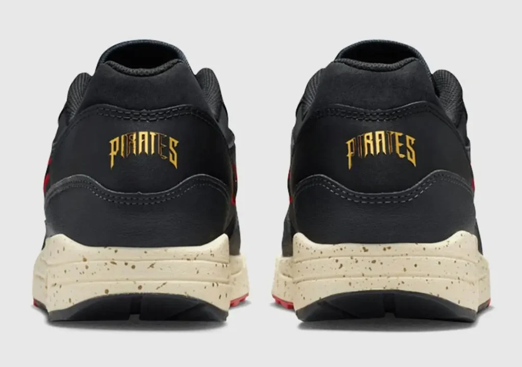 Rear view of a pair of black Nike Air Max 1 shoe featuring gold “PIRATES” lettering across the heel tabs. The shoes have smooth black leather uppers with clean stitching, a cream speckled midsole with a slightly aged finish, and subtle red accents visible along the outsole, set against a neutral background
