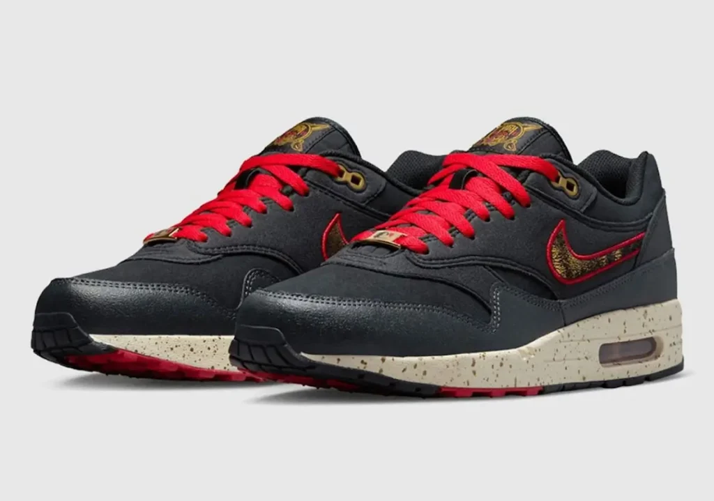 Angled front view of black Nike Air Max shoe with bold red laces and matching red-accented Swooshes featuring gold detailing. The upper appears in smooth black leather with subtle paneling, while the cream speckled midsole adds a vintage-inspired contrast. A visible Air unit sits in the heel, with touches of red on the outsole and gold eyelets and tongue branding completing the design against a neutral background