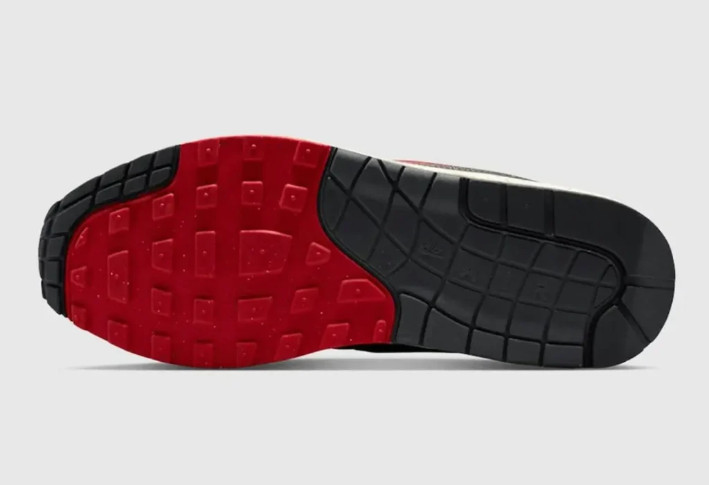 Bottom view of a Nike shoe outsole featuring a two-tone design with bold red rubber across the forefoot and black rubber at the heel. The tread pattern includes square and waffle-like textures for traction, with flex grooves running through the midfoot, set against a clean neutral background