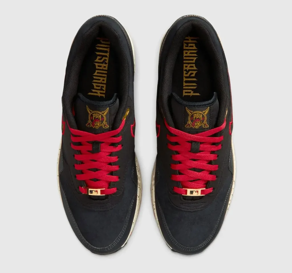 Top-down view of a pair of black Nike shoe featuring bold red laces and gold lace dubraes with a small logo detail. The tongues display a crest-style graphic in red and gold, while the insoles are printed with “PITTSBURGH” in a gothic-style font. The upper appears in smooth black material with subtle stitching, framed against a clean neutral background
