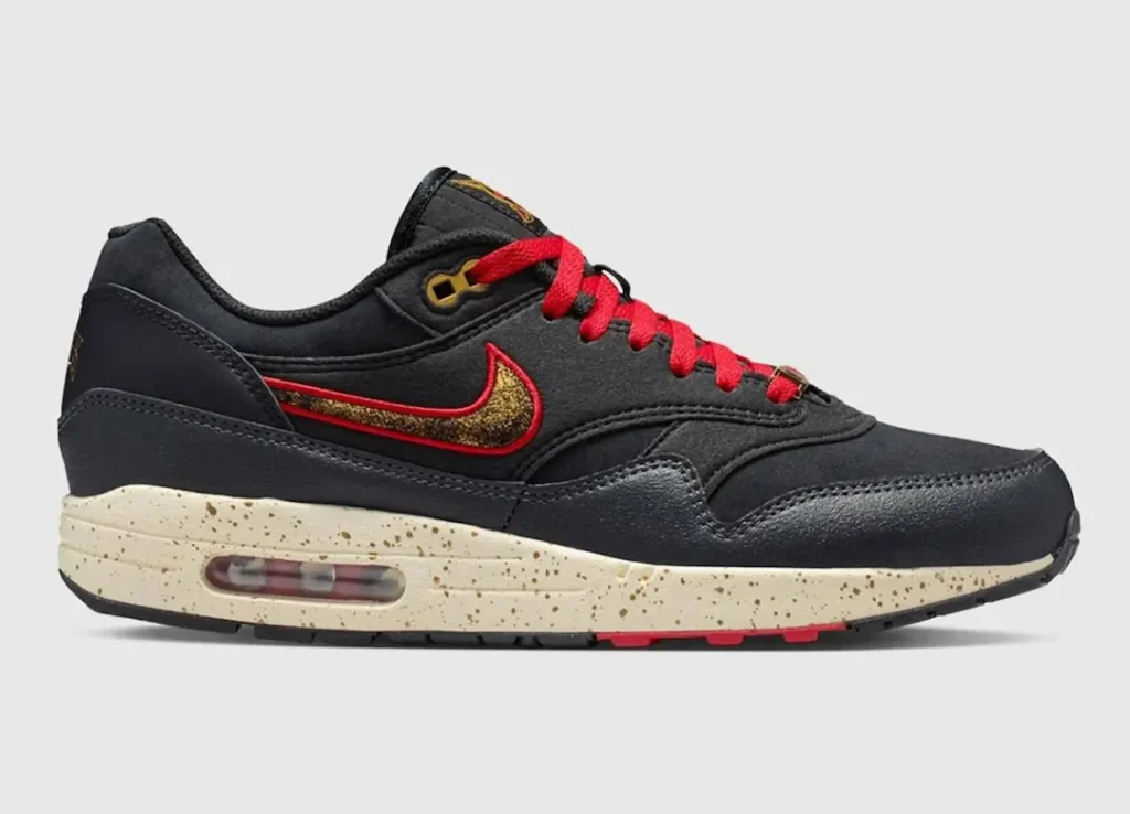Side profile of a black Nike Air Max shoe featuring red laces and a red-outlined Swoosh filled with gold detailing. The upper combines smooth and textured black materials with layered stitching, while the cream speckled midsole adds a vintage look and houses a visible Air unit in the heel. Subtle red accents appear on the outsole, and gold eyelets complete the design against a clean neutral background