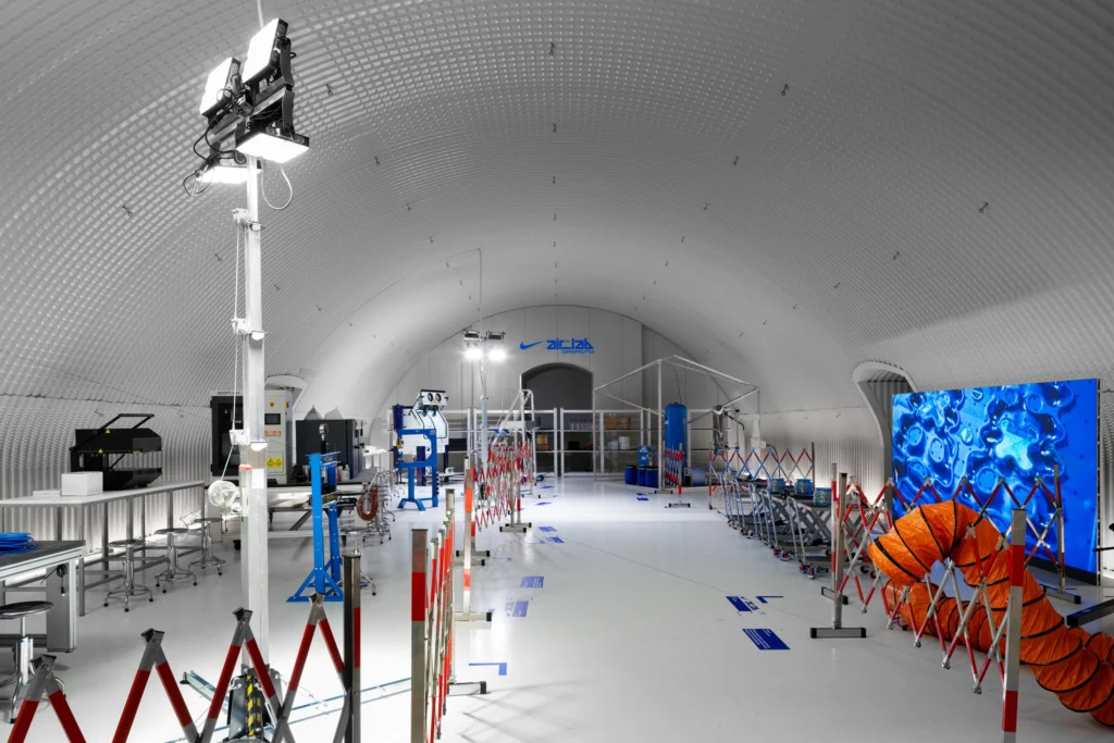 A wide-angle view inside a futuristic, tunnel-shaped laboratory space with a curved, ribbed white ceiling. The environment is brightly lit and highly controlled, featuring industrial testing equipment, metal workstations, and safety barriers arranged along a central walkway. On the right, a large digital screen displays abstract blue liquid-like view, contrasting with an orange ventilation tube nearby. At the far end, Nike Air Lab branding is visible above a doorway, reinforcing the space as a specialized research and innovation facility