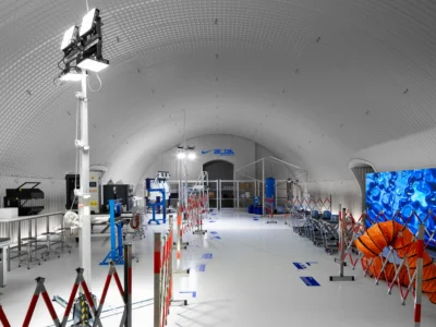 A wide-angle view inside a futuristic, tunnel-shaped laboratory space with a curved, ribbed white ceiling. The environment is brightly lit and highly controlled, featuring industrial testing equipment, metal workstations, and safety barriers arranged along a central walkway. On the right, a large digital screen displays abstract blue liquid-like view, contrasting with an orange ventilation tube nearby. At the far end, Nike Air Lab branding is visible above a doorway, reinforcing the space as a specialized research and innovation facility
