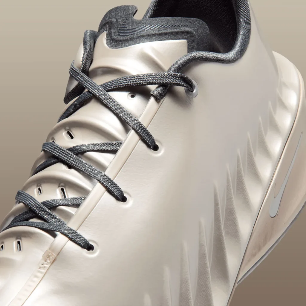 Close-up of a Nike basketball shoe upper in a champagne-beige finish, showing dark gray laces threaded through clean eyelets, a smooth molded surface with subtle ridged contours, and a partially visible Swoosh near the midfoot, highlighting a sleek, minimal performance design
