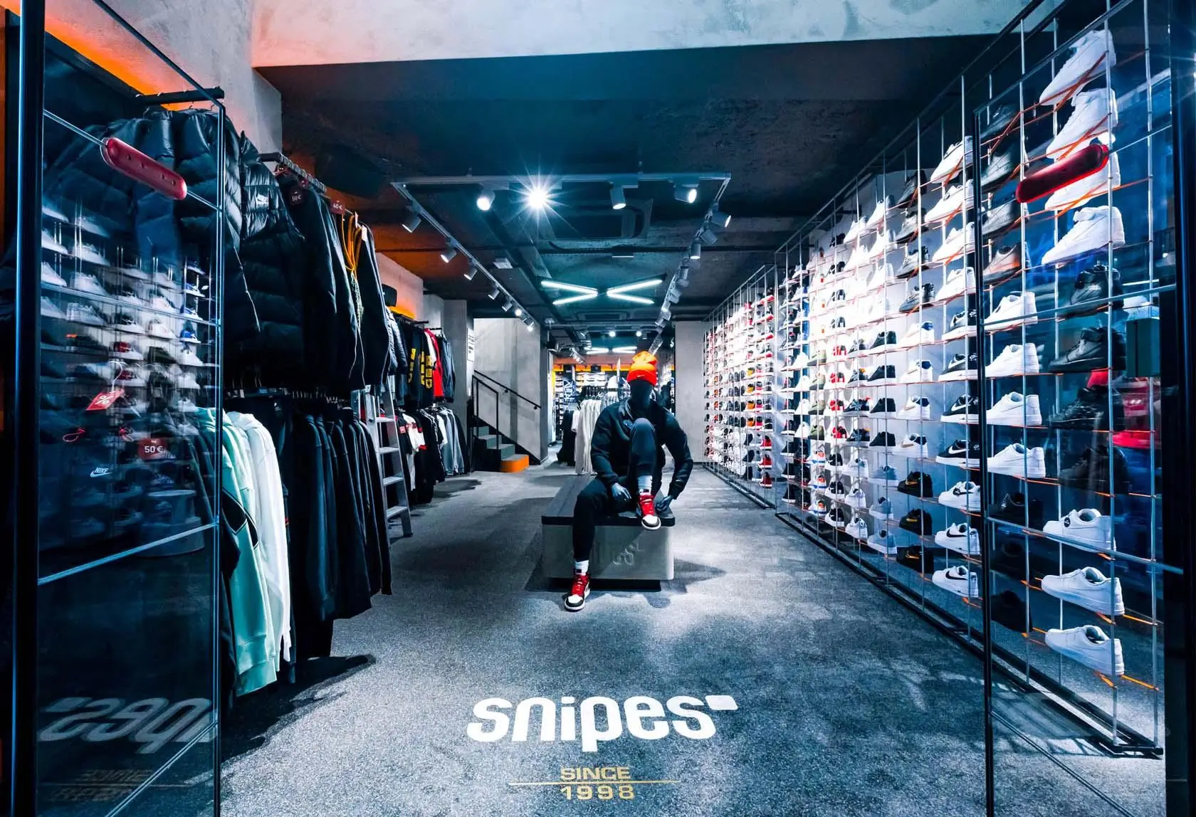 Modern Snipes retail store interior with sleek industrial design, featuring walls lined with rows of sneakers in glass displays, clothing racks with jackets and apparel on the left, and a person in a black outfit and orange beanie sitting on a central bench adjusting red sneakers under bright overhead lighting