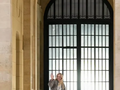 Wide, vertically framed scene centered on a grand architectural entryway with a tall, arched stone structure. The warm-toned stone walls and layered arch details create depth and symmetry, drawing attention toward a large black double door with grid-patterned glass panels that glow with diffused light from behind. In the foreground, a casually dressed person stands slightly off-center, mid-gesture, raising a hand in a friendly wave. They wear a relaxed, oversized plaid shirt layered over a light top, paired with loose dark trousers and casual footwear. A lanyard hangs from their neck, suggesting an event or backstage context. The scale contrast between the monumental doorway and the individual adds a sense of openness and atmosphere. Low ground lighting fixtures line the sides of the passage, while the cobblestone floor enhances the historic, European feel of the setting. The overall composition blends architectural grandeur with a candid, human moment.