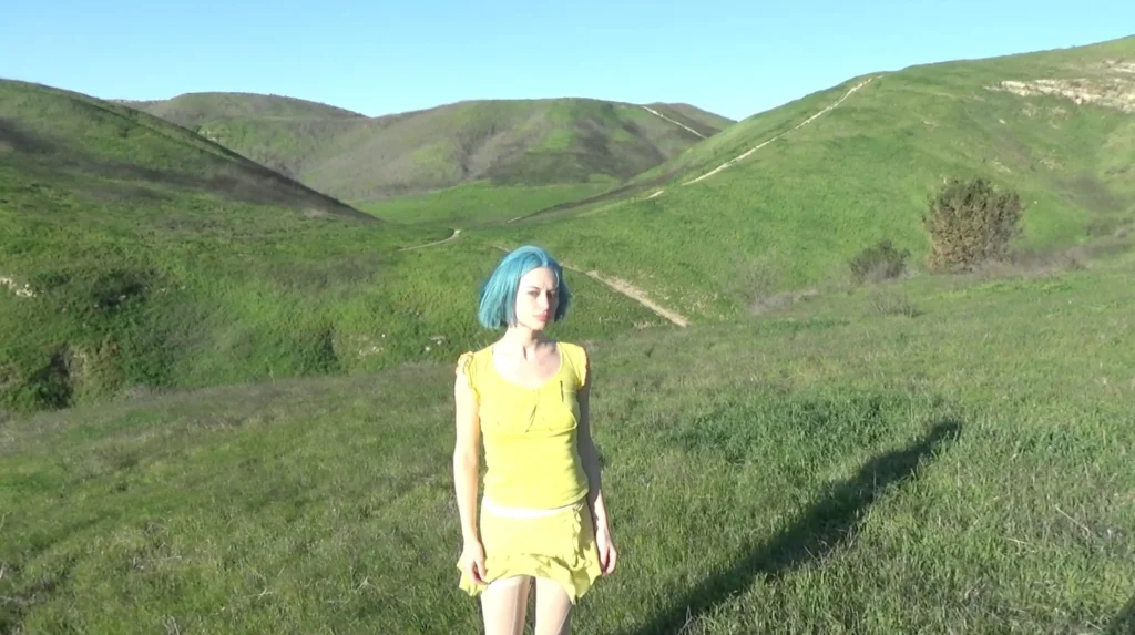Oxis with short blue hair stands in a sunlit expanse of rolling green hills, wearing a soft yellow dress that contrasts vividly against the landscape, evoking a quiet, introspective mood tied to nature and openness