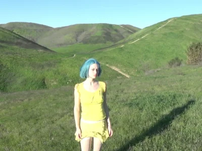 Oxis with short blue hair stands in a sunlit expanse of rolling green hills, wearing a soft yellow dress that contrasts vividly against the landscape, evoking a quiet, introspective mood tied to nature and openness