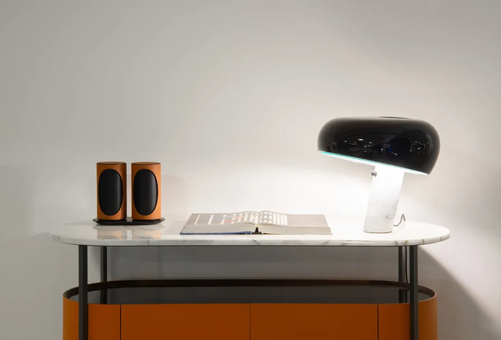 Pair of Geneva Lab Duo speakers in a warm orange finish set on a marble console table beside an open book and a sculptural black dome lamp, within a minimalist interior that balances modern design and soft ambient lighting
