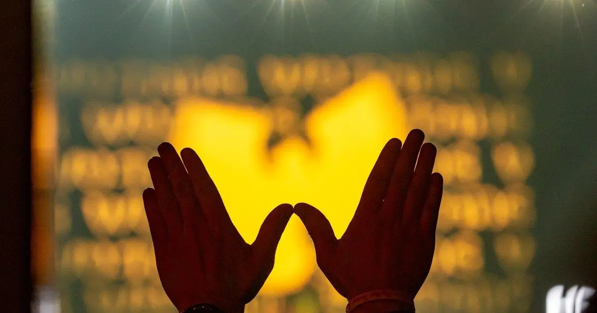 Silhouetted hands raised and forming the iconic Wu-Tang “W” symbol in front of a glowing yellow Wu-Tang logo backdrop, with blurred text behind suggesting a live event or performance setting