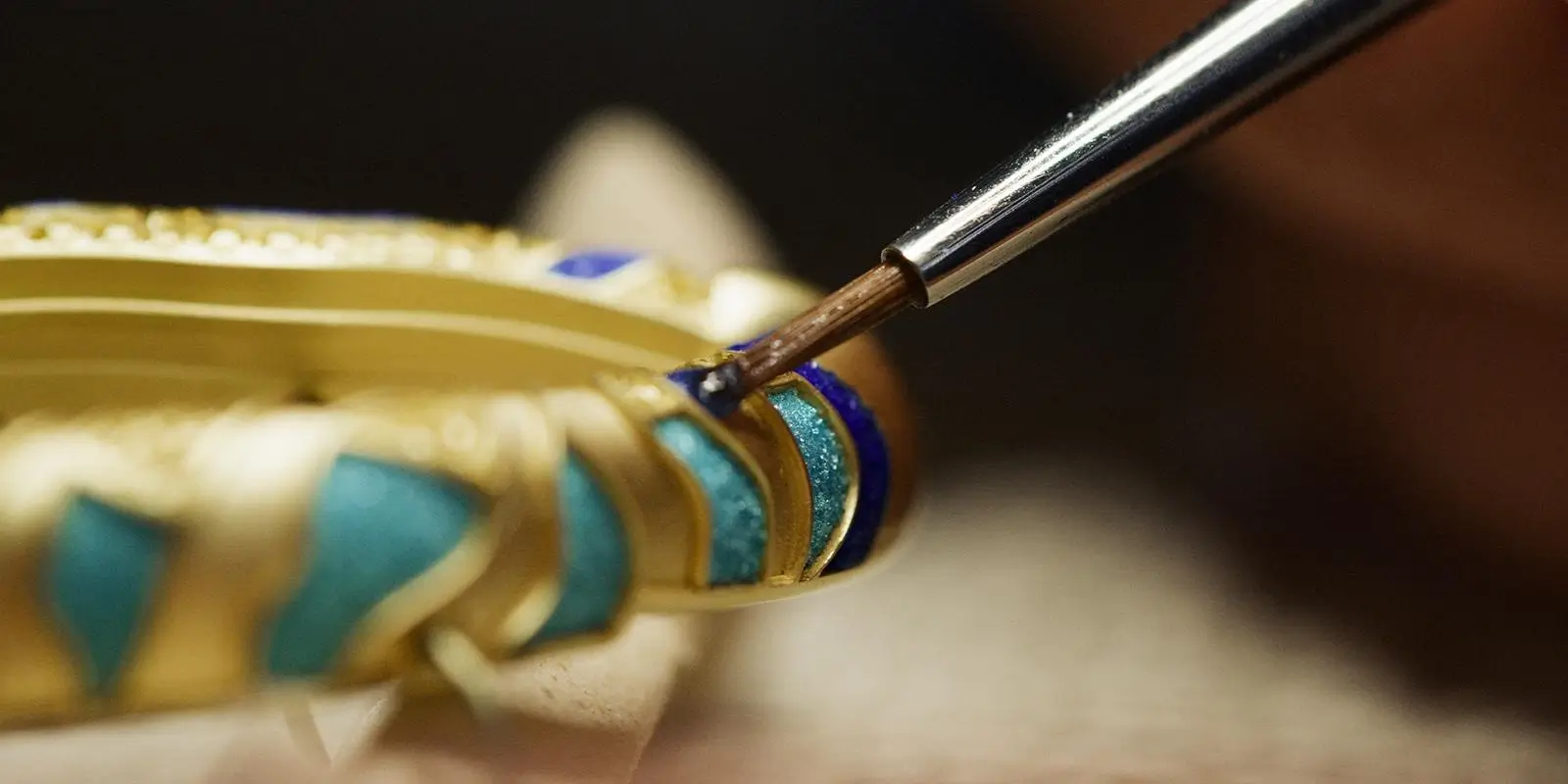 Fine brush applying deep blue enamel into recessed segments of a gold watch component, revealing champlevé technique in progress with precise, hand-controlled detailing