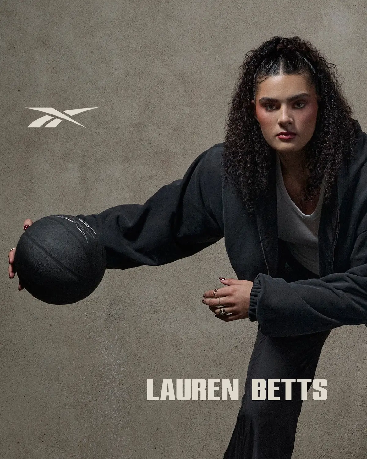 Portrait of Lauren Betts in a Reebok campaign, holding a basketball mid-motion while wearing a dark athletic outfit, set against a textured neutral background with Reebok logo and her name displayed