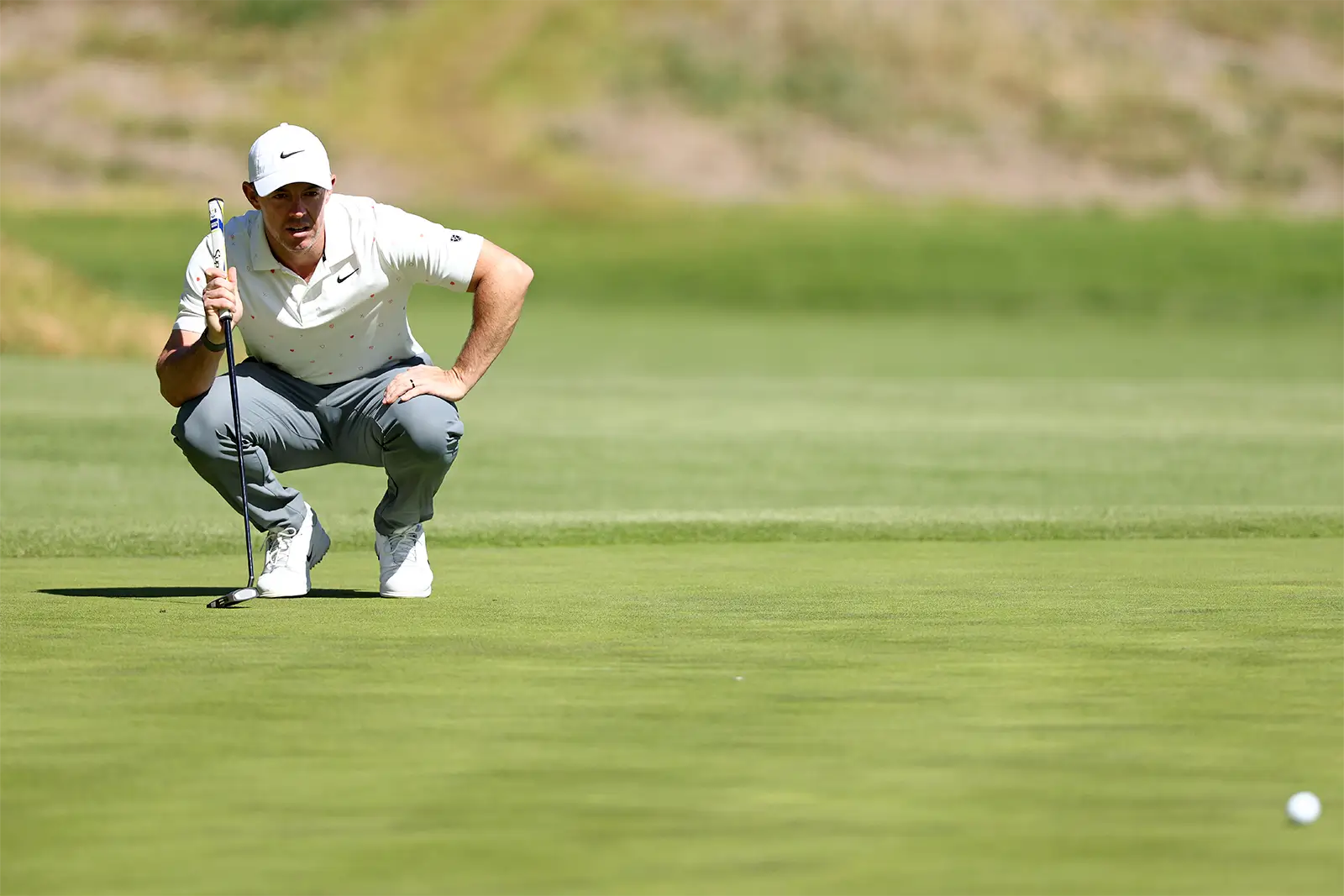 Rory McIlroy crouches low on a sunlit green, reading a putt with focused intensity, dressed in a white Nike cap and polo with gray trousers as the blurred fairway stretches behind him
