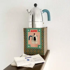 Alessi Vite espresso maker by Philippe Malouin displayed atop its original patterned box, featuring a polished aluminum body, teal thermoplastic handle, and black knob lid against a minimal interior backdrop