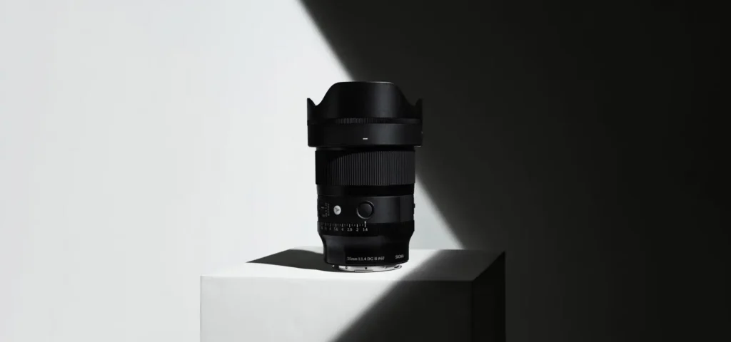 A black Sigma camera lens sits centered on a white pedestal, dramatically lit by a diagonal beam of light that splits the background into stark light and shadow, emphasizing its textured rings and sculptural form