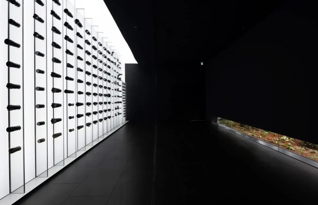 A long, minimalist corridor unfolds in sharp contrast: on the left, a luminous white wall stretches into the distance, punctuated by a precise grid of mounted camera lenses that protrude uniformly like sculptural instruments. Each unit is evenly spaced, creating a rhythmic, almost mechanical repetition—part gallery display, part industrial meditation. The lighting is clinical yet intentional, casting soft shadows that emphasize the cylindrical forms and their quiet precision