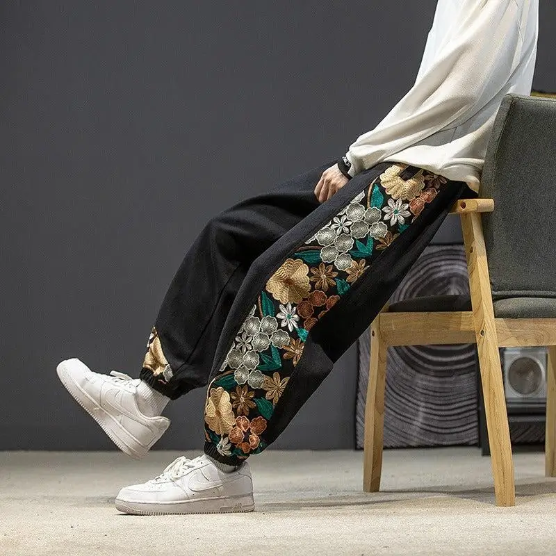 Black embroidered floral joggers with rich gold, silver, and teal detailing, paired with a white sweatshirt and white Air Force 1s. Elevated streetwear aesthetic