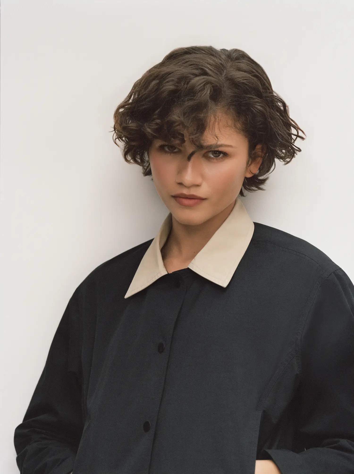 Zendaya in a close-up portrait wearing a minimalist black jacket with a contrasting beige collar, her short tousled curls framing a focused, composed expression against a neutral backdrop