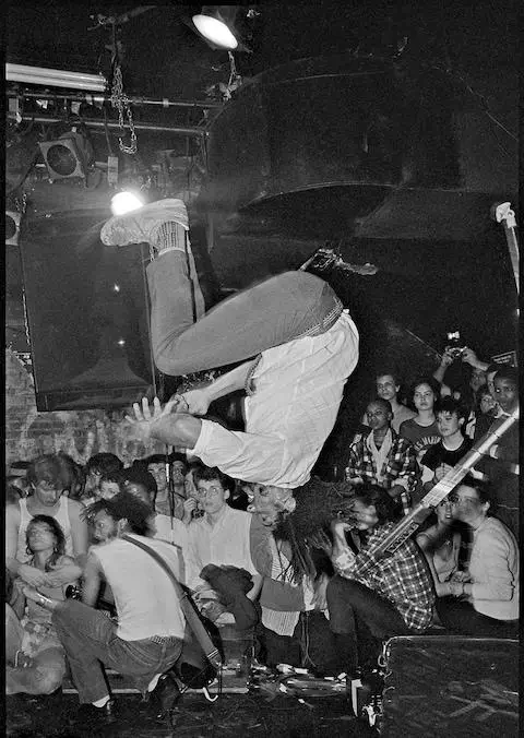 photograph capturing a chaotic live music moment inside a packed club. A performer is frozen mid-backflip above the stage, body fully inverted with legs tucked and arms out, while band members continue playing beneath him—one crouched low, another gripping a guitar to the right. The crowd presses in tightly around the stage, faces turned upward in anticipation, some reaching out. Overhead lights and exposed fixtures frame the scene, amplifying the raw, high-energy atmosphere of an underground punk or hardcore show