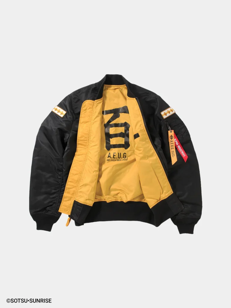 Close-up of a black MA-1 bomber jacket sleeve and chest area featuring embroidered gold patches reading “Alpha Industries” and “Q. Bajeena.” The sleeve includes a utility pocket with dual hanging tags—one red “Remove Before Flight” tag and one yellow A.E.U.G. tag—alongside a striped patch with three gold stars, all set against a smooth nylon fabric