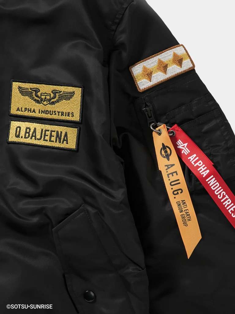 Black MA-1 bomber jacket shown from the front with a slightly glossy nylon finish and ribbed collar, cuffs, and hem. The design features gold embroidered chest patches reading “Alpha Industries” and “Q. Bajeena,” matching three-star sleeve patches on both arms, and a sleeve utility pocket with red “Remove Before Flight” and yellow A.E.U.G. tags, with a hint of yellow lining visible at the collar