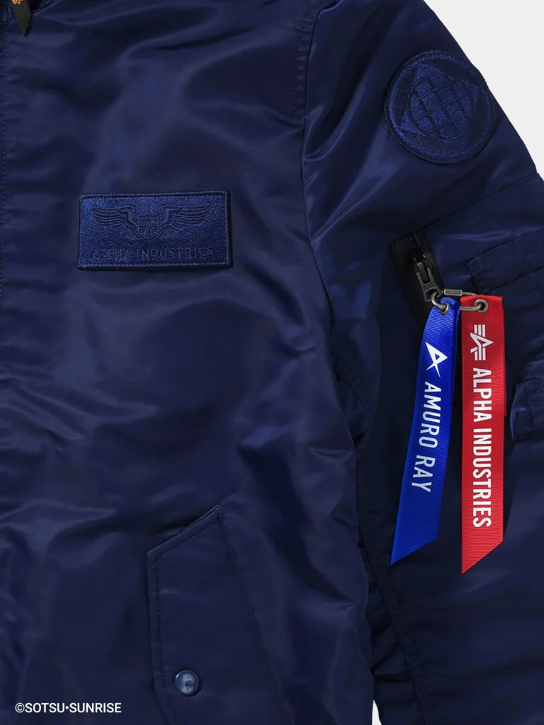 Close-up of a navy blue MA-1 bomber jacket showing a tonal “Alpha Industries” chest patch and a circular sleeve patch. The sleeve utility pocket features two hanging tags—a red “Remove Before Flight” tag and a blue tag reading “Amuro Ray”—set against smooth, slightly glossy nylon fabric