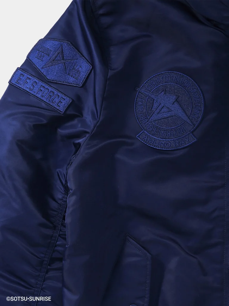 Close-up of a navy blue hooded MA-1 style bomber jacket showing tonal embroidered patches on the chest and sleeve. The sleeve features an “E.F.S.F. FORCE” patch and a geometric insignia, while the chest displays a circular emblem with “Amuro Ray,” all rendered in subtle monochromatic stitching on smooth, slightly glossy nylon fabric