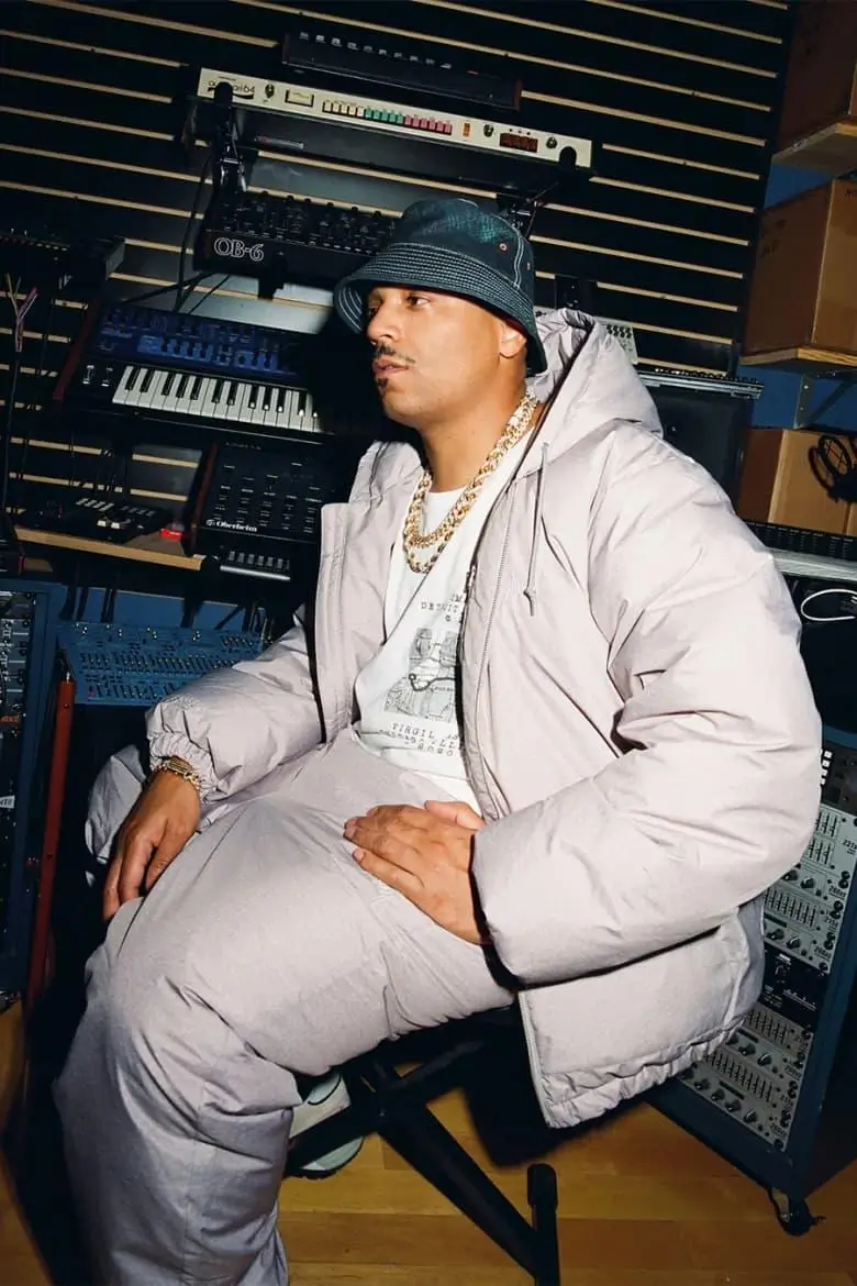 Artist in the studio rocking a full light gray puffer set, bucket hat, and gold chains surrounded by synths and recording equipment. Signature Skate Muzik vibe