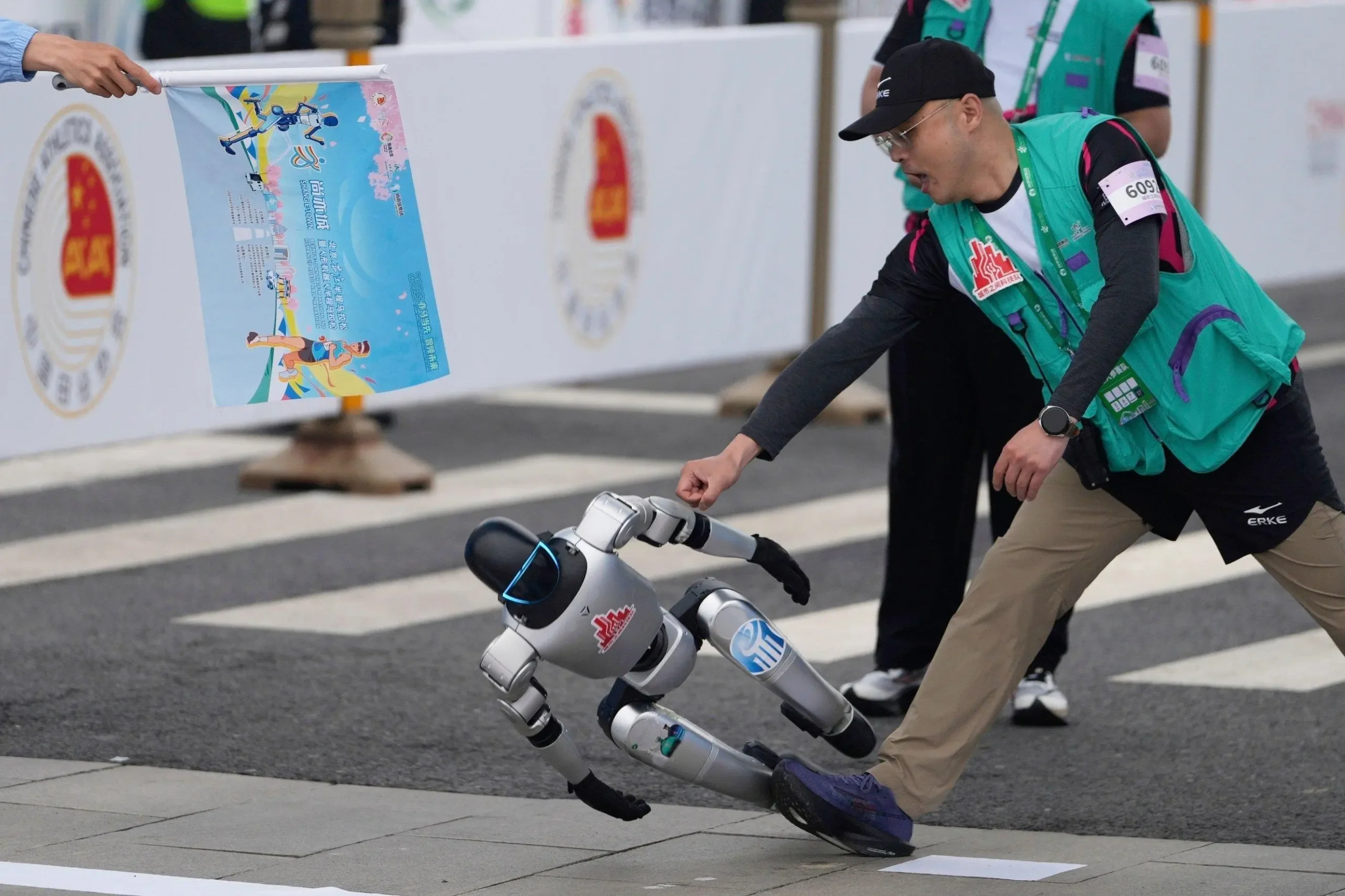 A humanoid robot falls sideways on a racecourse as an event staff member reaches out to assist during a public robotics running event