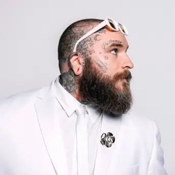 Teddy Swims in a studio portrait wearing a white suit and sunglasses on his head, showing facial tattoos and a full beard while looking to the side