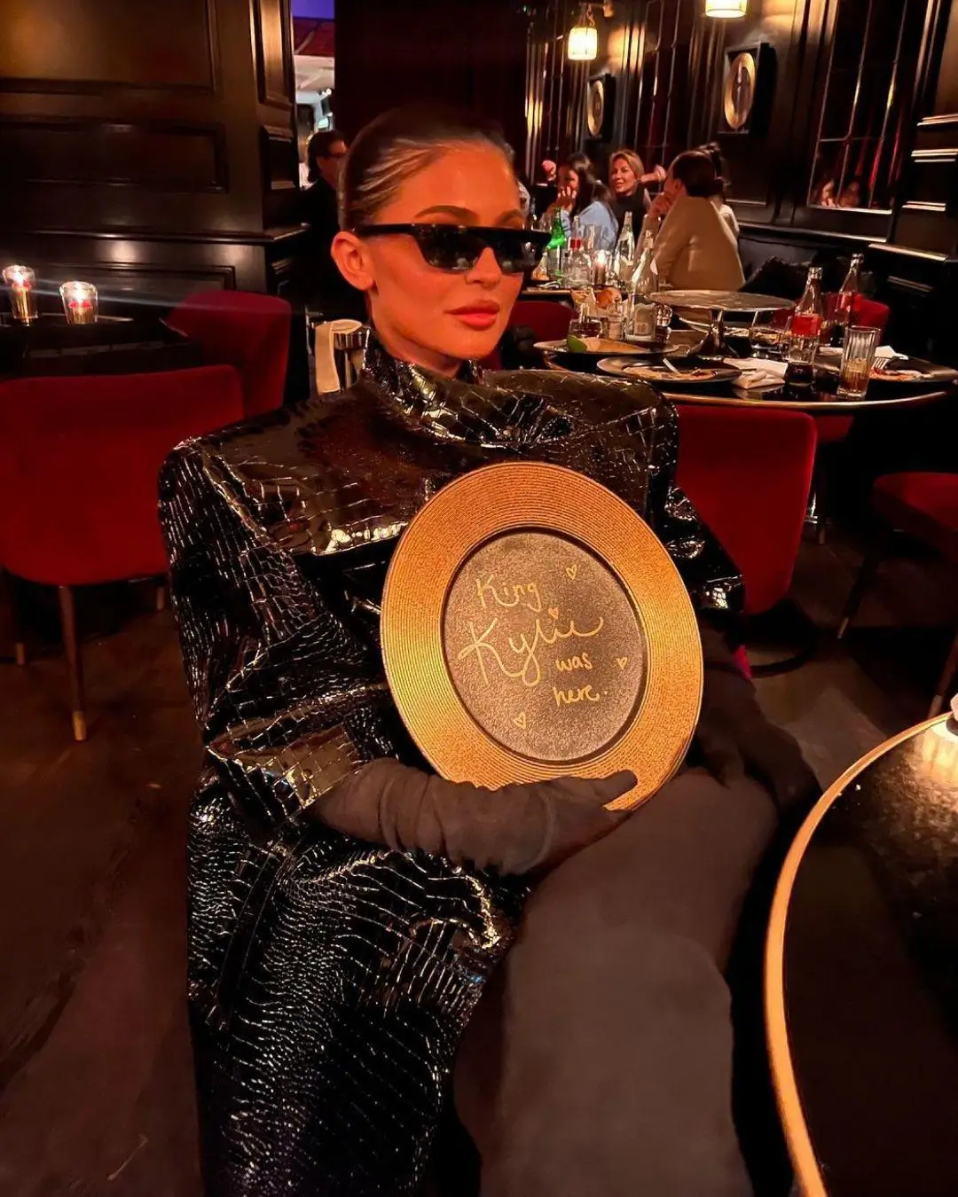 Elegant woman in a dimly lit restaurant, dressed in a glossy black textured outfit with matching gloves and dark sunglasses, holding a decorative plate inscribed “King Kylie was here,” seated among red velvet chairs and candlelit tables in a chic, upscale setting