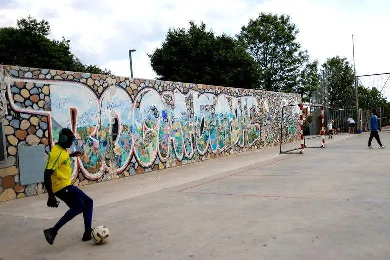 Person playing football on a concrete court beside a graffiti-covered wall, with a goal and others in the background