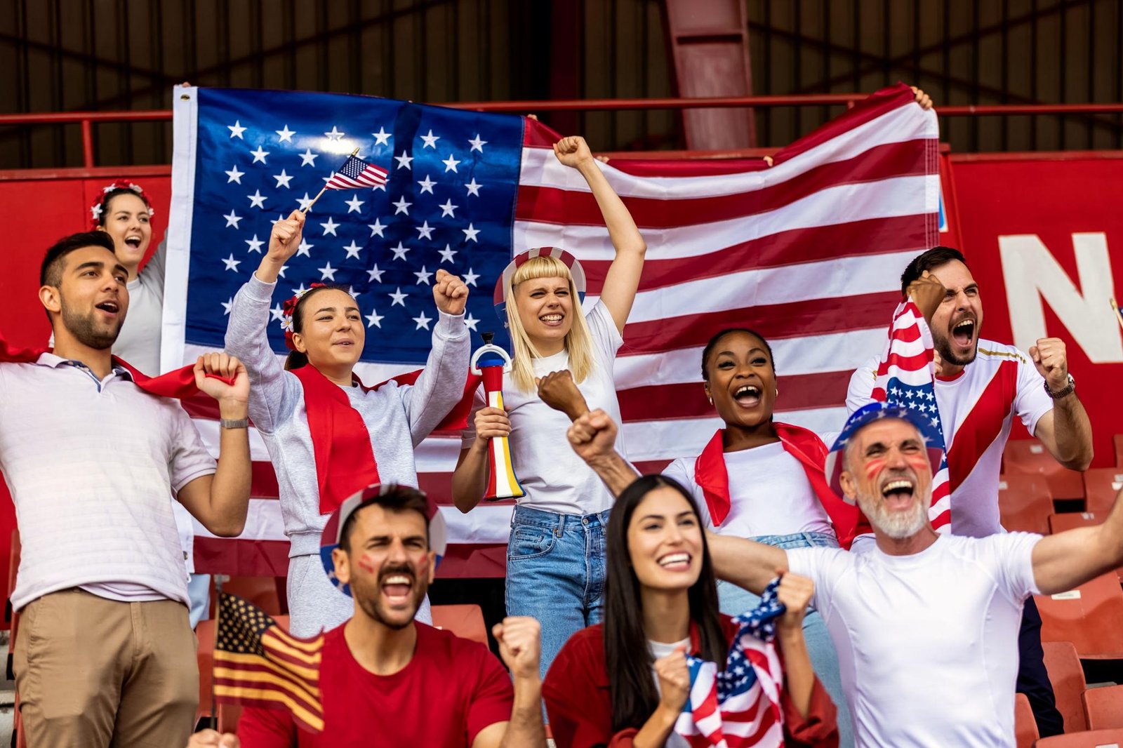 Cheering group of diverse fans in a stadium holding a large American flag, raising their fists and waving small flags in celebration during a lively sporting event
