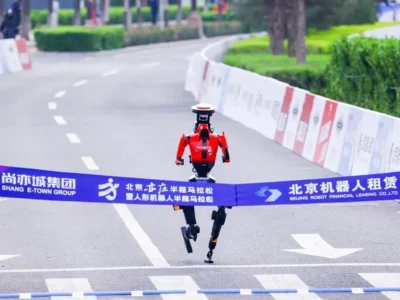 A red humanoid robot crosses a marathon finish line on a city street, breaking a branded ribbon as it runs along a barricaded course lined with trees and greenery