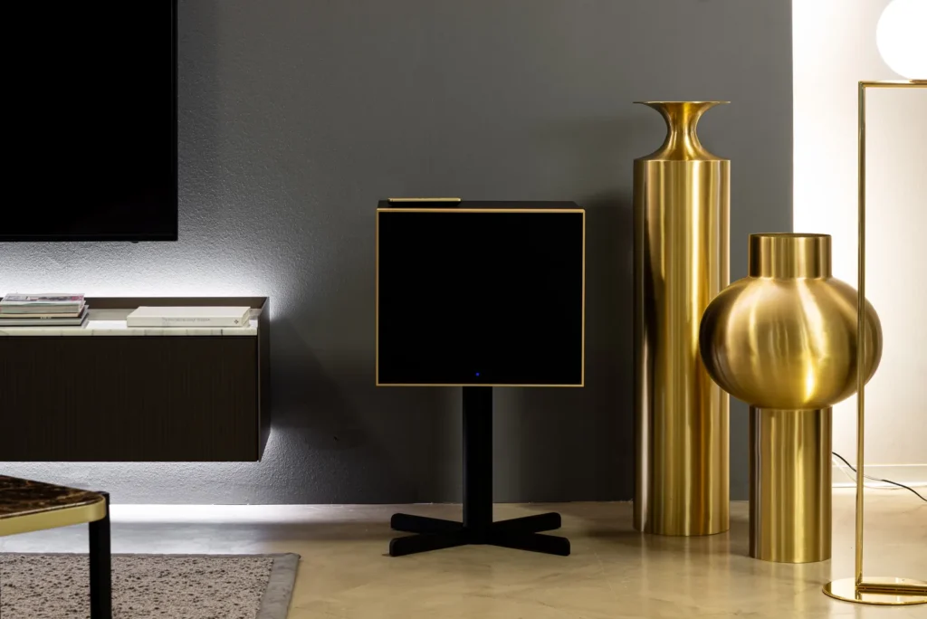 Geneva Lab DeCon/XL speaker in a black finish with gold trim on a minimalist stand, positioned in a refined interior beside sculptural gold vases and a floating media console against a dark accent wall