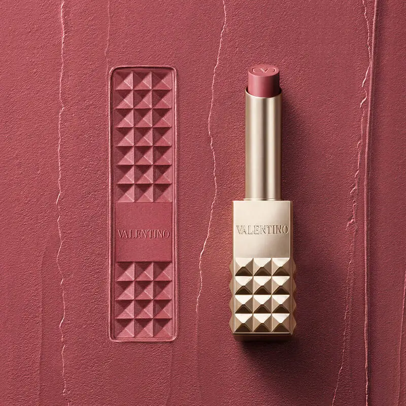 A close-up of a Valentino lipstick set against a textured dusty-rose surface, featuring a gold studded case with the brand name engraved and a matching pink-toned lipstick bullet, alongside a rectangular imprint of the signature stud pattern embossed into the background