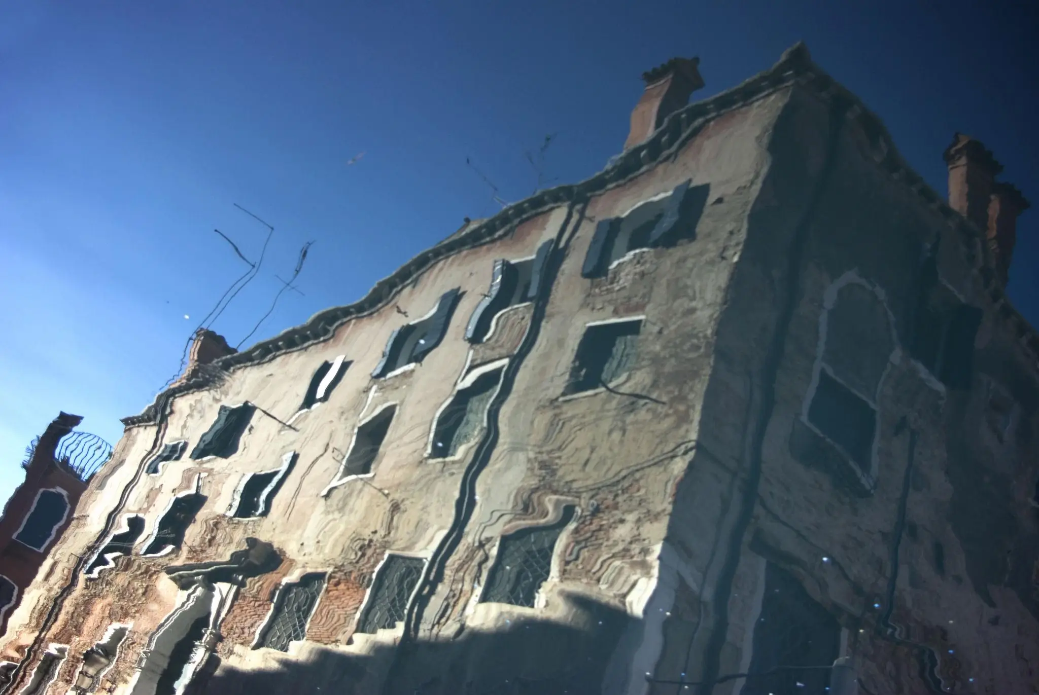 Historic building façade seen as a wavy reflection in water, with distorted windows and rooflines under a bright blue sky, creating an abstract, fluid visual effect