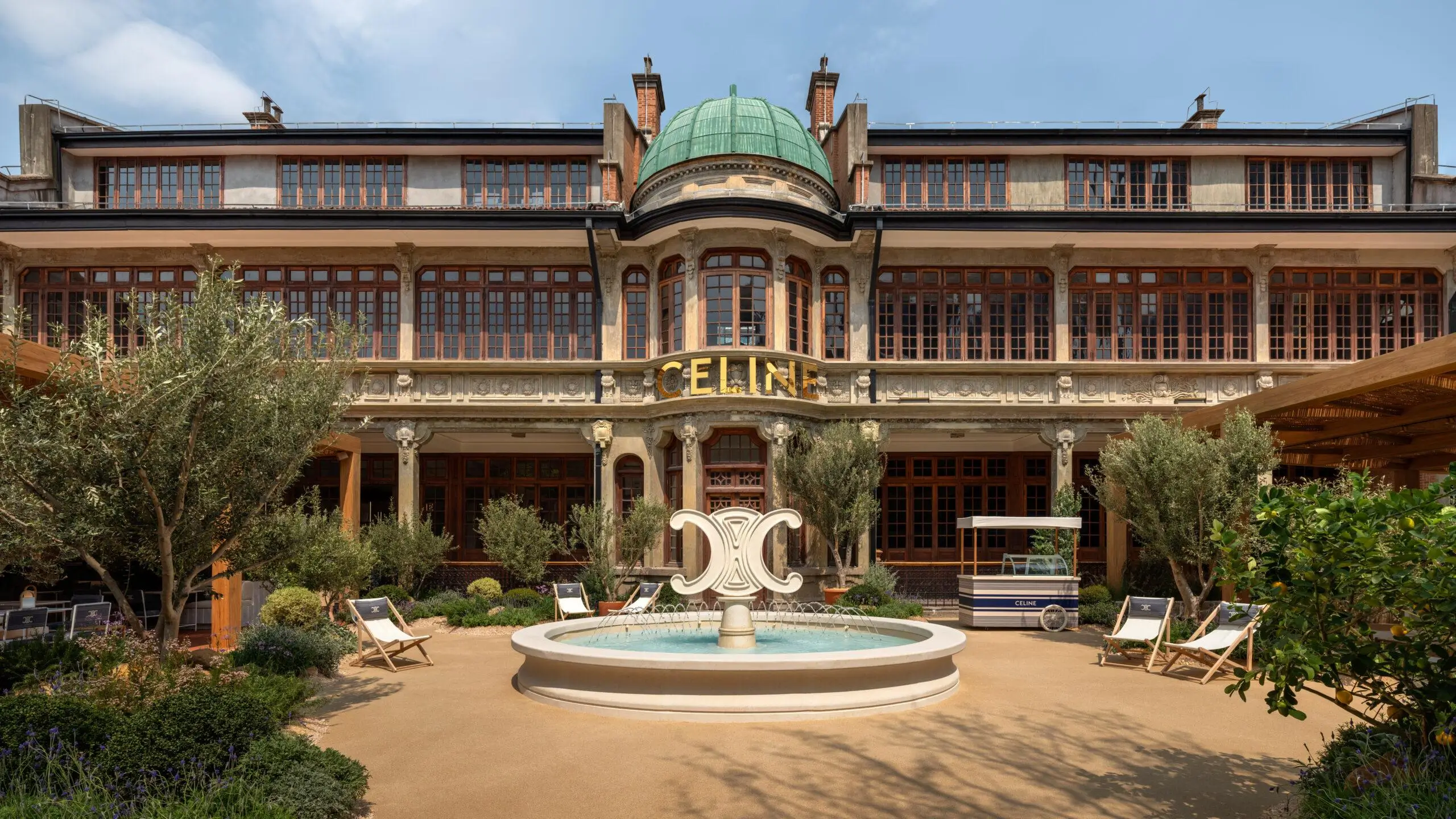 Historic Celine-branded building with ornate facade and green dome, centered around a courtyard fountain and landscaped garden with lounge chairs and a branded kiosk